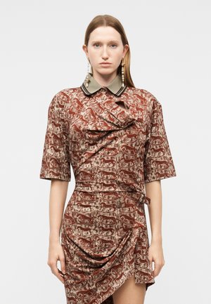 Žena v hnedom vzorovanom šate s motívmi psov, olivovo zeleným golierom a asymetrickým závesom, doplnenom dlhými náušnicami s perlami.