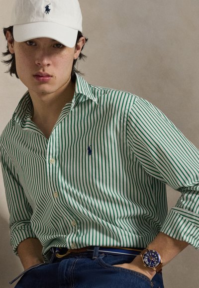 Jeune homme portant une casquette blanche et une chemise à rayures vertes à boutons avec manches retroussées, un jean en denim et une montre-bracelet à bracelet en cuir marron.