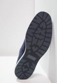 Chaussure en daim bleu avec une semelle en caoutchouc noir texturée, présentant un motif géométrique pour l'adhérence et une étiquette de taille 42 sur la semelle.