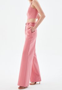 Woman wearing high-waisted wide-leg pink trousers with a matching cropped top and beige open-toe heels, hands in pockets, posing sideways.