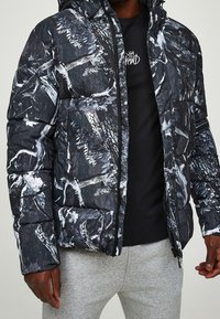 Muž v černo-bílém vzorovaném péřovém kabátu, černém tričku s grafikou a světle šedých teplákách, s rukama u těla.