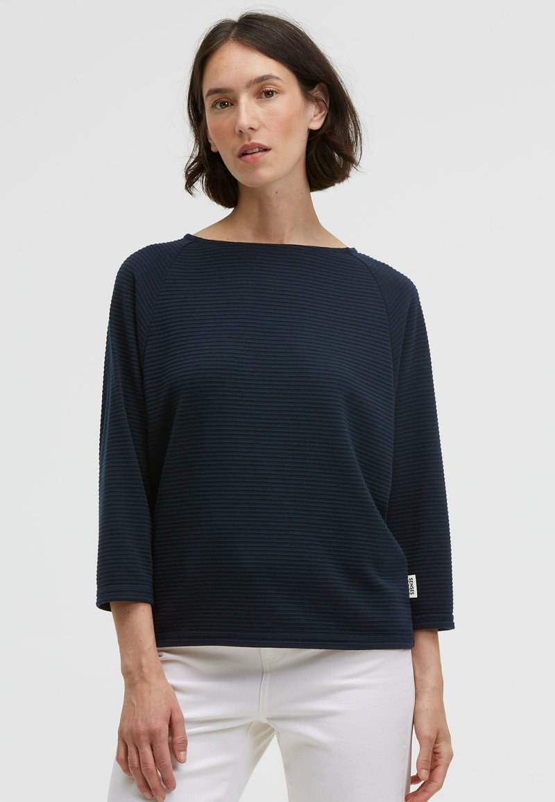 Marineblauer gerippter Pullover mit weitem Ausschnitt und Dreiviertelärmeln, kombiniert mit weißen Hosen. Besitzt ein kleines Etikett am Ärmel.