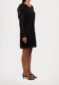 Svart blommig spetsklänning med långa ärmar, texturerat mönster och figurnära passform, matchad med glänsande svarta klacksandaler.