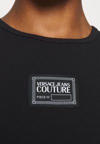 Svart bomullsskjorta med rund halsringning, som har en vit broderad etikett som säger "VERSACE JEANS COUTURE" och "PIECE N°."