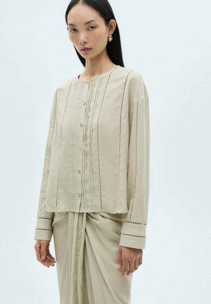 Light beige button-up shirt made of soft fabric, featuring vertical stripe detailing and cuffed sleeves. Drape style complements a skirt.