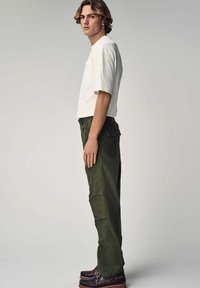 Chemise blanche à manches courtes, pantalon utilitaire vert olive, vue de côté. Le pantalon possède de grandes poches ; les chaussures sont marron foncé avec des coutures accentuées.