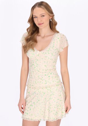 Unge kvinde med langt, bølget blondt hår iført en lys, blomsterprintet kjole med korte ærmer, stående mod en ensfarvet hvid baggrund.