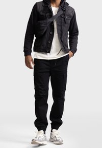 Man wearing black hooded denim jacket, white t-shirt, black cargo pants, white sneakers, and black crossbody bag, standing with hands in pockets.