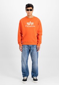 Felpa arancione con il logo bianco "Alpha Industries", abbinata a jeans blu e sneaker nere. La felpa presenta un colletto e polsini a coste.