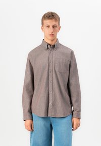 Joven con cabello corto, vistiendo una camisa de manga larga marrón abotonada y jeans azul claro, de pie contra un fondo blanco liso.