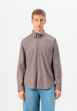 Young man with short hair wearing a buttoned-up brown long-sleeve shirt and light blue jeans, standing against a plain white background.