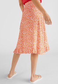 Bloemige wikkelrok in pastelroze met rode en gele bloemen, met een uitlopende zoom en lichte stof. G gedragen met witte sandalen.