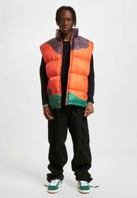Puf vest i orange og grøn med grå detaljer, der har høj krave, frontlynlås og kviltet design. Båret over en sort top.