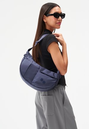 Frau trägt schwarze Sonnenbrille, ein schwarzes, kurzärmeliges Oberteil, graue Hose und trägt eine große marineblaue Umhängetasche über einer Schulter.