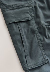 Jambe de pantalon cargo en tissu vert foncé avec coutures visibles et une poche à rabat rectangulaire avec une couture d'angle diagonale.