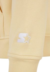 Manica di felpa gialla con polsino a coste, caratterizzata da un piccolo logo ricamato bianco di una lettera "S" stilizzata e una stella. Texture morbida.