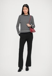 Maglione a righe bianco e nero, pantaloni neri su misura, stivaletti neri con tacco e una piccola borsa a tracolla rossa. Sfondo bianco semplice.
