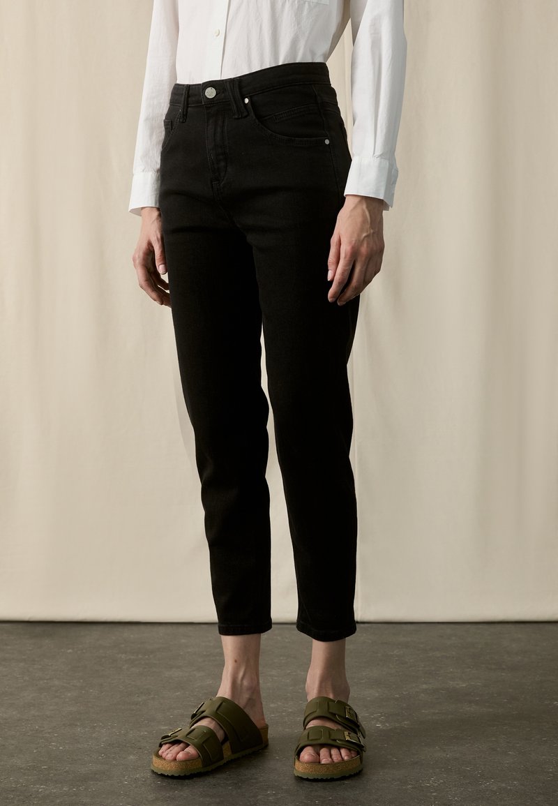 Personne portant un pantalon noir slim, une chemise blanche à manches longues et des sandales vert olive à double bride, debout sur un sol gris.