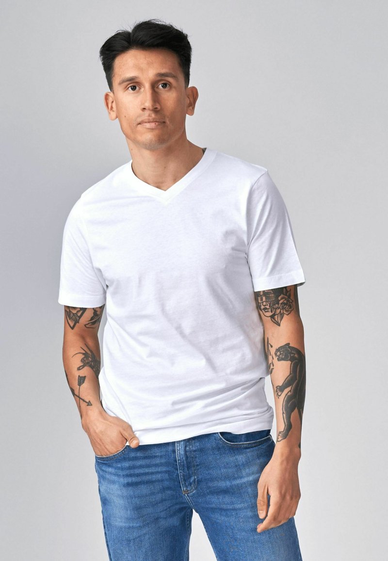 Homme aux cheveux courts noirs portant un T-shirt blanc à col en V et un jean bleu, montrant des tatouages sur les deux avant-bras, debout devant un fond gris uni.