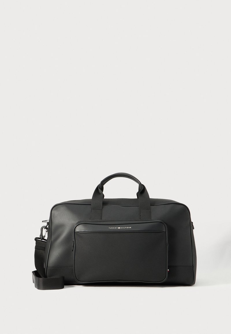 Sac de voyage noir en matériau synthétique lisse. Il dispose de deux poignées et d'une poche zippée à l'avant, avec une bandoulière amovible.