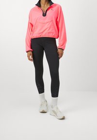 Neon pink, pitkähiha pullover, jossa on puolivetoketju ja etutasku, yhdistetty mustiin leggingseihin, joissa on teksturoitu muotoilu, ja valkoisiin sukkiin.