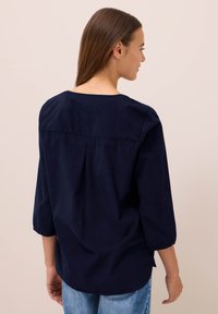 Vrouw met haar rug naar de kijker, gekleed in een marineblauwe blouse met driekwartmouwen en een blauwe spijkerbroek, tegen een effen achtergrond.