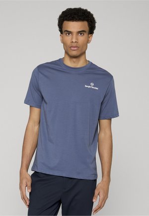 Junger Mann mit kurzen lockigen Haaren, der ein blaues Sergio Tacchini T-Shirt und dunkle Hosen vor einem schlichten hellgrauen Hintergrund trägt.