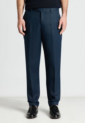 Homme portant un pantalon formel bleu marine et des mocassins en cuir noir, debout devant un fond clair uni.
