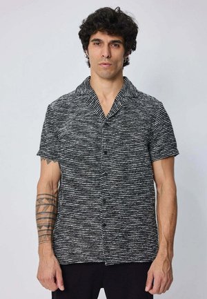 Homme aux cheveux bouclés foncés portant une chemise texturée à manches courtes rayée noir et blanc, debout devant un fond clair uni.