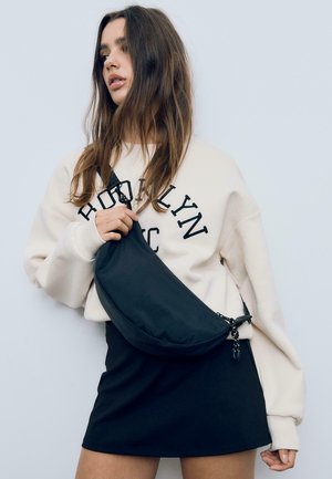 Borsa a tracolla nera realizzata in tessuto morbido, dalla forma curva con chiusura a zip, indossata sopra un pullover color crema e una gonna nera. Design minimalista.