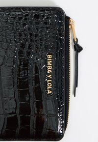 Portefeuille en faux cuir noir avec embossage crocodile, logo doré, accent de fermeture éclair doré, et finition texturée. Forme rectangulaire compacte.