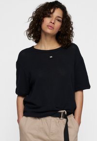 Top nero a maglia con scollatura rotonda e maniche corte, con un piccolo dettaglio del logo rettangolare sul davanti. Da abbinare a pantaloni beige.
