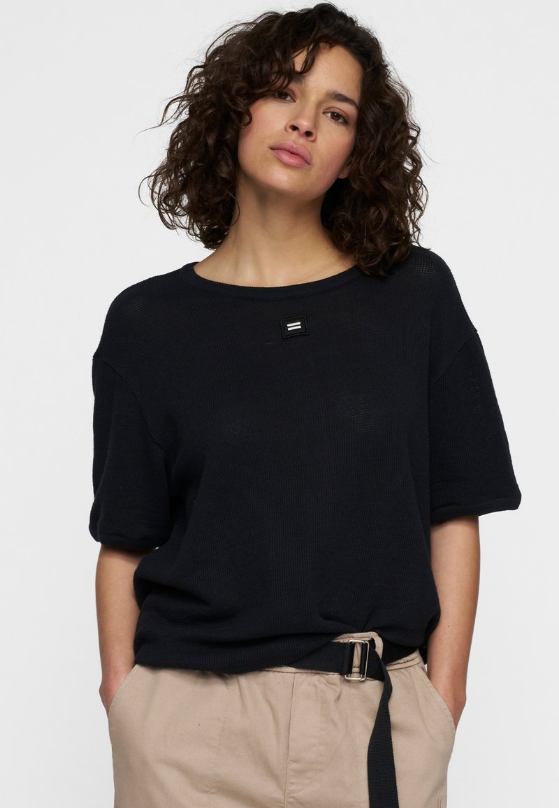 Top nero a maglia con scollatura rotonda e maniche corte, con un piccolo dettaglio del logo rettangolare sul davanti. Da abbinare a pantaloni beige.
