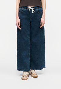 Άτομο που φοράει άνετο σκούρο μπλε τζιν, παντελόνι με φαρδιά πόδια και λευκό κορδόνι, με μπεζ αθλητικά παπούτσια με ζωικά σχέδια.