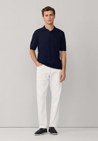 Hombre de pie contra una pared gris, vistiendo un polo azul marino de manga corta, pantalones blancos con una mano en el bolsillo, y zapatos oscuros sin cordones.