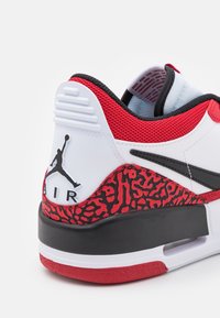 Vit och röd sneakers med en texturerad svart hälpanel, med en svart Swoosh och en Jumpman-logotyp. Inkluderar röda accenter och meshelement.