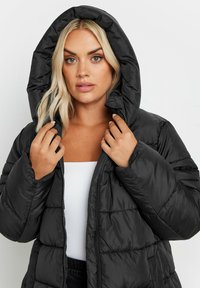 Svart puffjacka med huva, som har en kviltad textur och framdragkedja. Tyget har en slät yta och ett lätt utseende.