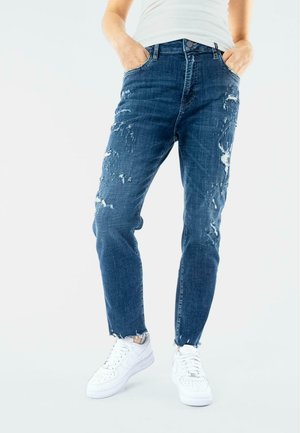 Jeans in denim blu con dettagli strappati, gamba affusolata, design a cinque tasche e orlo sfilacciato; indossati con sneakers bianche.