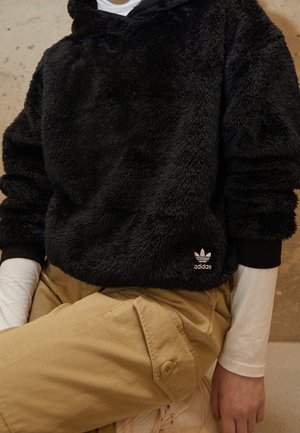 Osoba ubrana w czarną, puszystą bluzę z kapturem Adidas na białej koszuli z długim rękawem i beżowych spodniach z zasuwaną na guziki kieszenią, siedzi.