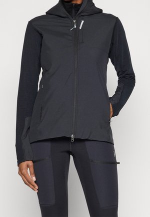 Persona che indossa una giacca nera con cappuccio e zip con tasche e leggings neri con tasche con cerniera sulle cosce, mani rilassate ai lati.