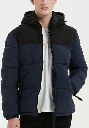 Doudoune avec un corps bleu marine, des épaules noires et une capuche. Elle est équipée d'une fermeture éclair, de poignets élastiques et d'une texture matelassée.
