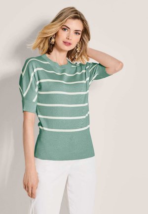 Blonde Frau mit schulterlangem Haar trägt grünen Kurzarmpullover mit weißen Streifen und weißen Hosen, posiert vor einer beigen Wand.