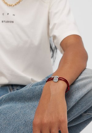 Rotes Schnurarmband mit einem runden Silberanhänger, der das Wort "STUDIOS" trägt. Das Armband wird mit einem kleinen silbernen Toggle-Verschluss gesichert.