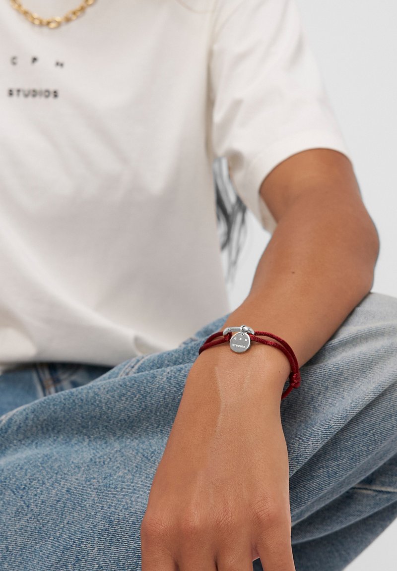 Bracciale di corda rossa con un charm rotondo d'argento con la parola "STUDIOS". Bracciale fissato con un piccolo fermaglio a Toggle d'argento.
