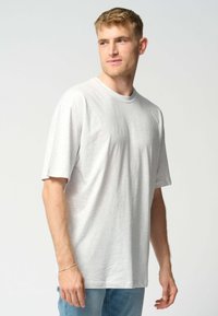 Ung mand iført en simpel hvid oversized T-shirt og lyseblå jeans, som ser ud til siden mod en ensfarvet baggrund.