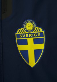 Escudo de futebol da Suécia com fundo azul, uma cruz amarela e o texto "SVERIGE" em negrito, colocado sobre um tecido escuro.