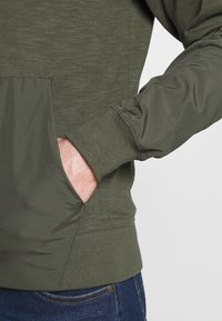 Olivově zelená mikina s texturovanou horní částí a hladkými rukávy. Má přední kapsu a žebrované manžety.