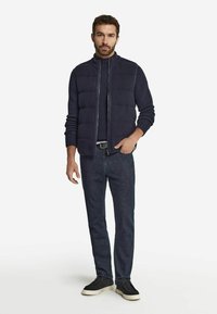 Marinblå stickad väst med hög krage och horisontella kviltade sektioner, kombinerad med mörka jeans och svarta skor med ljusa sulor.