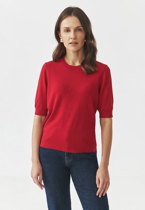 Rode gebreide sweater met korte mouwen, geribbelde halslijn en zoom. Gecombineerd met blauwe spijkerbroek, staat het model tegen een neutrale achtergrond.