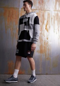 Svetlo šedá a čierna farba blokovaná kapucňová mikina s predným vreckom, skombinovaná s čiernymi šortkami s grafickým dizajnom a logom a so svetlo šedými atletickými topánkami.